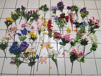 Full view of assorted artificial flowers arranged on tiled floor showing variety in color, type and size.
