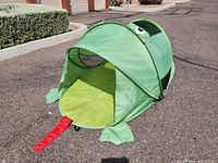 Green frog-shaped pop up tent set up outdoors, showing side angle with the tent entrance open and red tongue detail on the floor outside.
