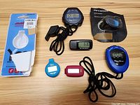 Lot of assorted pedometers and stopwatches arranged on wood surface; includes devices both unpackaged and one unopened in packaging.