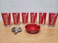 Six Coca Cola glasses with red and white Coca Cola logo and text, arranged in a row, small red Coca Cola ashtray in front, metal bottle opener beside ashtray.