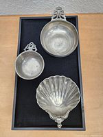 Photo showing all three pewter items arranged on a black tray: large bowl with ornate handle, small bowl with handle, and shell-shaped dish.