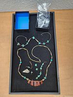 Top view of the tray containing two beaded necklaces, turquoise earrings, rings, a brooch, and a pin arranged on a black surface inside a box.