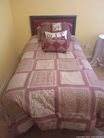 Photo of twin bed with red fabric upholstered headboard and bed with brown patterned quilt and pillows in a yellow room with hardwood floor.