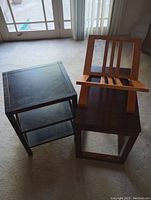 Two small wooden end tables and one wooden magazine rack shown on floor. End tables are square with dark finish, one has two shelves, the magazine rack has slatted sides and top holder.