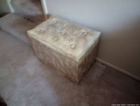 Front angle view of the small tan storage ottoman showing cushioned top with metal ring pull handles; fabric heavily weathered.