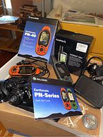 Photo showing DeLorme Earthmate PN-40 GPS device with box, quick start guide, and Bushnell binoculars on top of a cardboard box with various items in background.