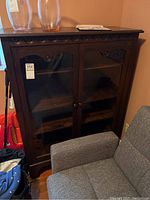 Front view of the bookcase showing clear glass doors and adjustable shelves inside in a tight corner next to grey upholstered furniture with vases on top.