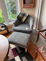 Photo showing gray tub-style chair with curved back and arms, wooden legs, placed near window with matching ottoman and plaid throw on chair back.