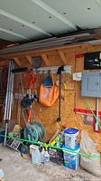 Garage wall with hanging garden tools, hose reel, hose, clothesline, orange gardening bag, and various supplies on floor