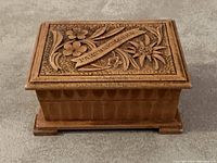 Closed view of the rectangular antique wooden jewelry box showing the lid with detailed floral carvings and engraved text 'l'Artenkikchen'. The box has carved border patterns along the edges and a natural wood finish.