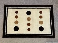 Front of rectangular bathroom floor rug mat with unique circle patterns in black, brown, beige on cream background and double border lines in black and beige.