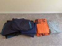 Stack of 17 assorted heavy quilted, lightweight Uhaul, and light blue textile moving blankets against a beige wall on carpeted floor.
