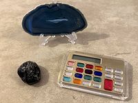 Photo of all three items together: polished blue quartz agate slice displayed on a clear stand, a black polished rock, and a gemstone calculator with colorful gemstone buttons on a grey surface.