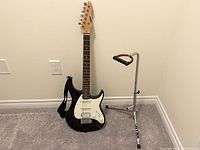 Full view of black Peavey Raptor Plus electric guitar with strap attached and metal stand placed beside it on carpeted floor against white wall.