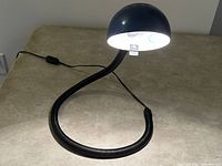 Blue enameled shade lamp lit on a table with flexible black snake-style neck coiled and bent in a curve shape showing multidirectional adjustability