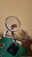 Metal hands-free magnifier stand showing lens and adjustable clamps on a black weighted base, sitting on top of the Red Cross First Aid book.