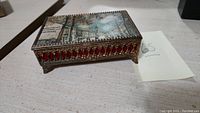 Closed view of metal music box showing silk street scene on lid and red inset decoration on sides.