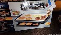 Box image showing griddle with pancakes and bacon cooking on top, and warming tray below. Displays product branding and features.