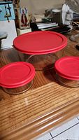 Three Pyrex clear glass bowls of varying sizes each topped with a red lid, placed on a wooden surface.