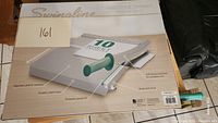 Box of Swinline guillotine paper cutter with visible features such as alignment grid, protective guard, green handle, and noted 10 sheet capacity.