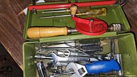 Green metal toolbox with a red hand crank drill, wooden handle screwdriver, wrenches, and various small tools inside.