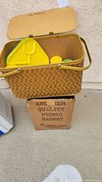 Large woven picnic basket sitting on original box, showing wooden panel lid and two carry handles.