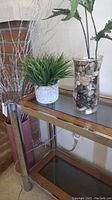 View of the two-shelf wood and glass table with chrome legs showing faux green plant in white pot and tall faux plant in glass vase with stones
