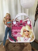 Barbie doll posed next to Barbie handbag case on wooden surface with light background