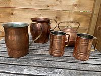 Overall view of all five brass items including two water pitchers, two cups, and one teapot on a wooden surface with wood panel background