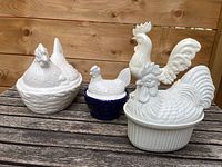 Photo of three ceramic covered dishes shaped like chickens and roosters showing detailed sculpting and glossy white finish with one blue base dish.