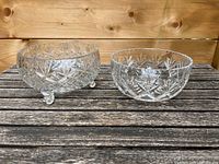 Two clear cut crystal bowls displayed on a wooden surface, one elevated on three small spherical feet and the other resting flat. Both bowls feature intricate cut designs including star and diamond patterns.