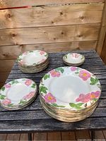 Group view of all plates and bowls stacked on a wooden table showing the distinctive pink rose and green leaf design on cream-colored ceramic.