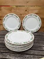 Stack of eight white dinner plates with holly and berry Christmas border and visible gold rim on a wooden surface.