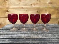 Front view of four red glass goblets with clear stems and bases placed on a wooden surface against a wood background.