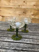 Front view of five clear dessert or champagne glasses with green stems and bases arranged on a wooden surface.