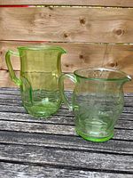 Two green glass water pitchers side by side on wooden surface with wood plank background. One pitcher taller and plain, other shorter with floral etching.