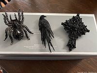 All three jewelry items displayed on a white box: spider ring, black bird brooch, and black cross brooch.