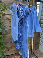 Photo showing five ladies clothing items hanging on hangers outdoors against a wooden fence: sleeveless white lace dress/nightgown, two blue and white striped dresses, light blue long-sleeved blouse, and a blue and white vertically striped top.