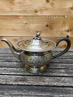 Full view of silver plated teapot showing embossed floral and scroll design on body, curved spout, and lid with decorative finial.
