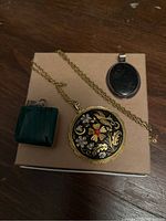 Photo showing two pendants and a gold tone chain necklace arranged on a cardboard surface, highlighting details of the floral pendant, the green rectangular stone pendant, and the oval black stone pendant.