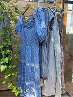 Four summer dresses hanging on wooden hangers outdoors with greenery and wood wall background, showing colors and styles