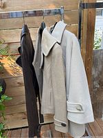 Three ladies coats hanging on rack outdoors: brown leather, beige wool, and light grey vintage Aquascutum wool coat.