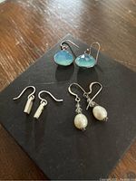 Photo of three pairs of earrings on a black box, including blue faceted stone drops, white pearl drops, and silver bar dangles