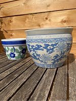 Two painted flower pots on wooden surface, larger blue and white floral pot in front, smaller blue, green and white floral pot behind