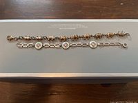 Two bracelets placed parallel on a white rectangular surface, showing one with elephant charms and the other with floral medallions.