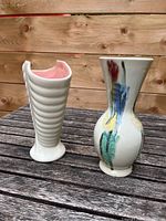 Photo showing two ceramic vases on a wooden surface. The vase on the left has a ribbed white exterior and pink interior. The vase on the right has a hand-painted floral design.