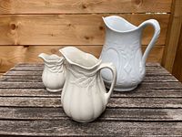 Three white ironstone jugs on a wooden surface, showcasing sizes and embossed details