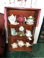 Front view of wooden three-shelf bookcase, showing decorative carved panels on sides and slightly raised top edge. Positioned against wall with various decorative items on shelves (not included).