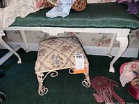 Full view showing the painted cast iron bench with dark green cushioned top and the floral tapestry footstool beneath it placed on green carpeting.