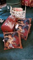 Photo showing white cast iron magazine rack on green carpet holding magazines, three angel-themed throw pillows with red and blue tones, and red hand painted wooden chest.